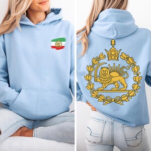May include: Light blue hoodie with a front pocket and a hood. The front features a small emblem of a flag with red, white, and green colors. The back displays a golden emblem of a lion holding a sword, surrounded by a wreath.