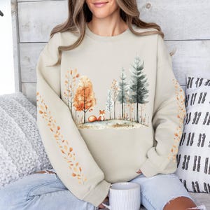 Pode incluir: Camisola bege com uma cena de floresta em aquarela com uma raposa, &aacute;rvores e folhas de outono. As mangas t&ecirc;m um desenho de folhas. A camisola &eacute; de cor neutra e tem um corte folgado.