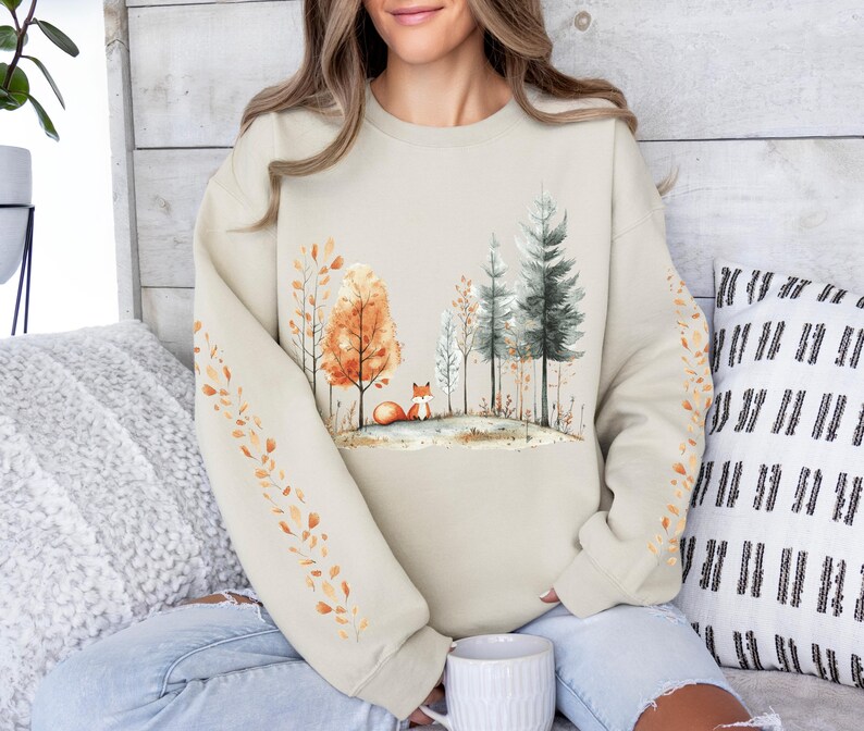 Pode incluir: Camisola bege com uma cena de floresta em aquarela com uma raposa, &aacute;rvores e folhas de outono. As mangas t&ecirc;m um desenho de folhas. A camisola &eacute; de cor neutra e tem um corte folgado.