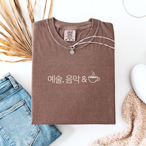 Peut inclure: Un t-shirt marron avec du texte cor&eacute;en et un dessin de tasse &agrave; caf&eacute;, associ&eacute; &agrave; un collier argent&eacute;, un jean bleu et des baskets blanches. Le t-shirt est pli&eacute; sur une surface blanche, avec une plante s&eacute;ch&eacute;e et une couverture beige.
