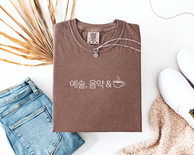 Peut inclure: Un t-shirt marron avec du texte cor&eacute;en et un dessin de tasse &agrave; caf&eacute;, associ&eacute; &agrave; un collier argent&eacute;, un jean bleu et des baskets blanches. Le t-shirt est pli&eacute; sur une surface blanche, avec une plante s&eacute;ch&eacute;e et une couverture beige.