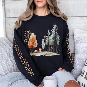 Pode incluir: Um moletom azul marinho com uma cena de floresta em aquarela com &aacute;rvores, uma raposa e folhagem de outono. As mangas t&ecirc;m um padr&atilde;o repetido de folhas douradas. O moletom &eacute; um estilo confort&aacute;vel e casual.