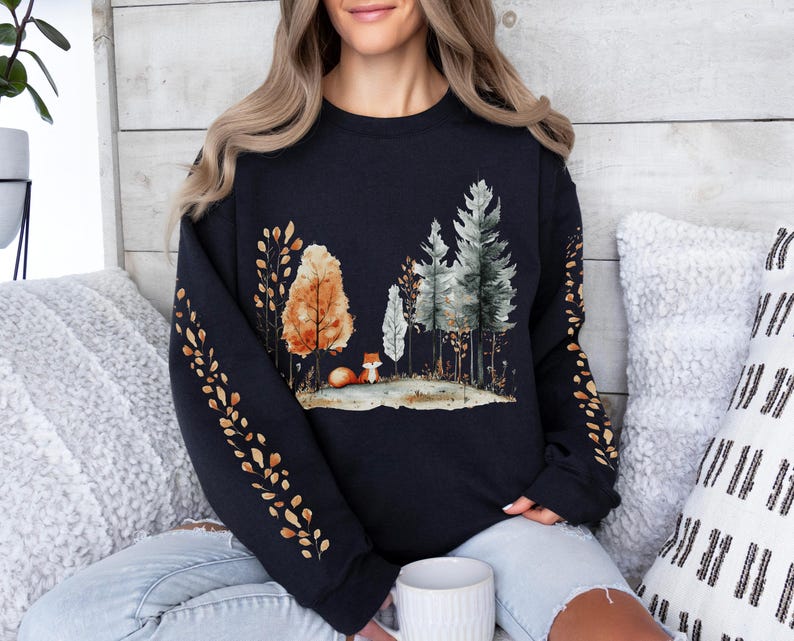 Pode incluir: Um moletom azul marinho com uma cena de floresta em aquarela com &aacute;rvores, uma raposa e folhagem de outono. As mangas t&ecirc;m um padr&atilde;o repetido de folhas douradas. O moletom &eacute; um estilo confort&aacute;vel e casual.