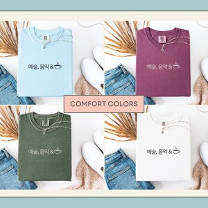 Peut inclure: Quatre t-shirts de diff&eacute;rentes couleurs : bleu clair, violet, vert olive et blanc. Chaque t-shirt porte le texte "예술, 음악 &" et un dessin de tasse &agrave; caf&eacute;. Les t-shirts sont pr&eacute;sent&eacute;s avec un jean, des chaussures blanches et un collier.