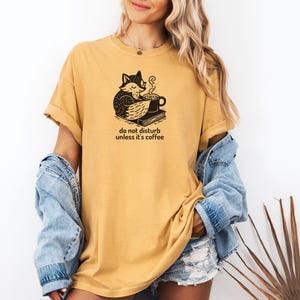 May include: Mustard yellow t-shirt with a black graphic of a fox, coffee cup, and book. The text "do not disturb unless it's coffee" is printed below the graphic. The shirt has short sleeves and a relaxed fit.