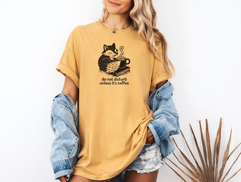 May include: Mustard yellow t-shirt with a black graphic of a fox, coffee cup, and book. The text "do not disturb unless it's coffee" is printed below the graphic. The shirt has short sleeves and a relaxed fit.