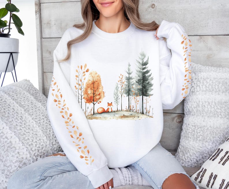 Pode incluir: Camisola branca com uma ilustra&ccedil;&atilde;o em aquarela de uma raposa em uma cena de floresta com &aacute;rvores laranja e verdes. As mangas t&ecirc;m um padr&atilde;o repetido de folhas laranja. O design evoca um tema de outono.