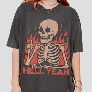 May include: Dark gray t-shirt featuring a skeleton giving a thumbs-up gesture, set against a backdrop of red flames. The phrase "HELL YEAH" is printed in red block letters below the skeleton. The shirt has a vintage, worn look.