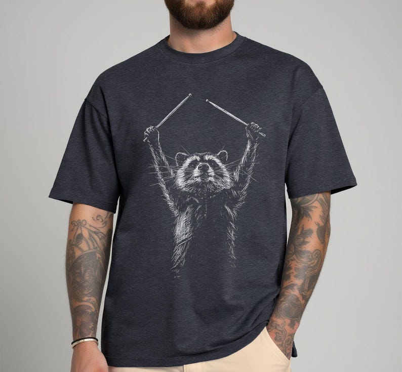 A man wearing a grey shirt featuring a detailed illustration of a raccoon holding drumsticks in the air. The casual, stylish design pairs well with his tattoos and denim.