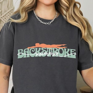 Peut inclure: T-shirt gris foncé avec l'inscription "BACKSTROKE swimmer" dans une police stylisée. Au-dessus du texte, un dessin d'un nageur orange. Le t-shirt est de style décontracté.