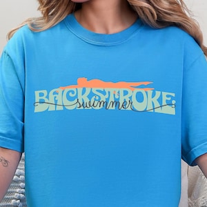 May include: A turquoise blue t-shirt with the words "BACKSTROKE swimmer" in a stylized font. Above the text is an orange illustration of a person swimming on their back. The shirt is made of a soft material.