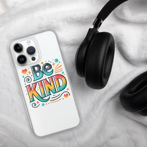 May include: A clear phone case with a colorful design that says "Be Kind". The case is on a white fuzzy surface next to black headphones.