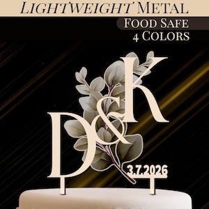 May include: A lightweight metal cake topper featuring the letters "D & K" and the date "3.7.2026". The design incorporates a floral element with green and brown leaves. The text "FOOD SAFE" and "4 COLORS" are also visible.