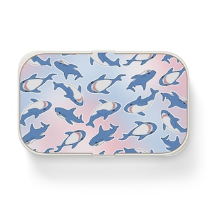 May include: A white rectangular lunchbox with a blue and pink ombre design featuring a repeating pattern of blue sharks with white teeth.