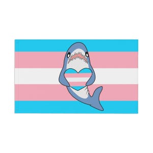 May include: A flag featuring the transgender pride colors of light blue, pink, and white stripes. A cartoon shark with a heart-shaped cutout is centered on the flag. The shark is light blue with a white belly and open mouth.