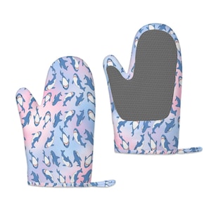 May include: Two oven mitts with a shark design. The mitts display a repeating pattern of blue sharks against a pastel pink, blue, and purple backdrop. One mitt's interior is grey, and both have a small loop for hanging, perfect for the kitchen.