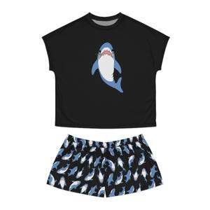 Op de afbeelding: Zwarte pyjamaset met een haaienontwerp. De top is zwart met een blauw-witte haaiengrafiek. De shorts hebben een herhalend patroon van blauw-witte haaien op een zwarte achtergrond. Een comfortabele pyjamaset.