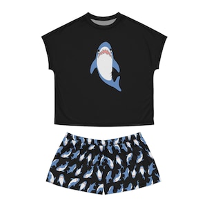 Könnte beinhalten: Ein schwarzes Kurzarm-Oberteil mit einer zentralen Grafik eines blau-weißen Hais mit offenem Maul. Die passende schwarze Shorts ist mit einem sich wiederholenden Muster aus kleineren blau-weißen Haien in verschiedenen Schwimmpositionen bedeckt.