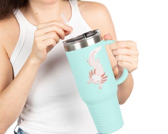 Puede incluir: Un vaso térmico azul claro con asa y tapa. El vaso presenta una ilustración de ajolote rosa. Está diseñado para bebidas frías o calientes e incluye una pajita reutilizable.