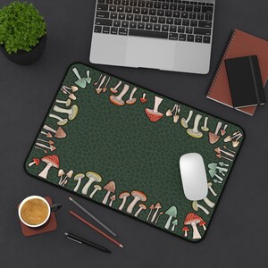 May include: A rectangular mousepad with a dark green background and a border of colorful mushroom illustrations. A white computer mouse sits on the pad, with a laptop, notebooks, and a cup of coffee nearby.