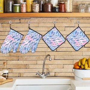 May include: A collection of kitchen textiles, comprising two oven gloves and two pot holders. They display a repeating pattern of blue and white whales against a pastel pink and blue backdrop. Each item has a black border and a hanging loop.