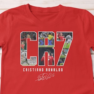 May include: A red t-shirt featuring the text "CR7" with images of Cristiano Ronaldo. The name "Cristiano Ronaldo" is printed below the text, along with his signature. The shirt is made of a soft material.