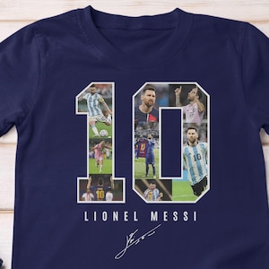 May include: Navy blue t-shirt featuring the number "10" composed of images of Lionel Messi. The name "LIONEL MESSI" is printed below the number, along with a signature. The shirt is displayed on a light wood surface.