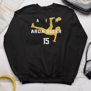 May include: Black sweatshirt featuring a yellow graphic of a soccer player in mid-air, with the word "AIR" above and "ARDA GÜLER 15" below. The design is centered on the chest.