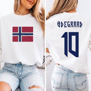 May include: White sweatshirt featuring the Norwegian flag on the front. The back displays the name "ØDEGAARD" and the number "10" in navy blue. The sweatshirt is a casual, sporty style.