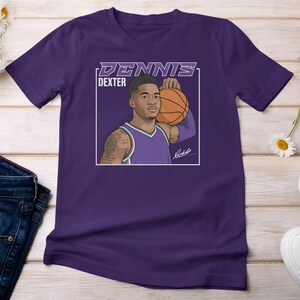 May include: A purple t-shirt featuring a graphic of a basketball player holding a basketball. The graphic includes the name "DENNIS DEXTER" in white and purple text. The player is wearing a purple jersey.