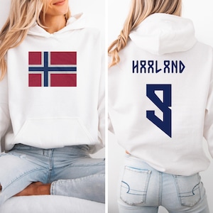 A woman wearing a white hoodie with the flag of Norway and the number "HRLAND" printed on the back.