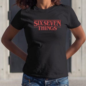 May include: Black t-shirt with the red text "SIXSEVEN THINGS" in a stylized font. The shirt is a crew neck and appears to be made of a soft material. The text is centered on the chest.