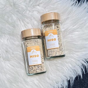 May include: Two glass jars with bamboo lids filled with a light brown powder. The jars have labels that say "Miso Mushroom Seasoning 2.8 oz (80g)".