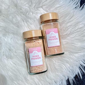 May include: Two glass jars with wooden lids filled with pink Himalayan salt and pink oyster mushroom powder. The labels on the jars read "Pink Power" and "Nourish Your Mushrooms".
