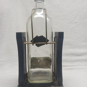 May include: A clear glass bottle with a black cap is held in a wooden stand. The stand is black with a gold-colored metal frame around the bottle. The bottle has a rectangular shape and a label.