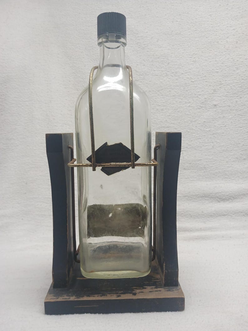 May include: A clear glass bottle with a black cap is held in a wooden stand. The stand is black with a gold-colored metal frame around the bottle. The bottle has a rectangular shape and a label.