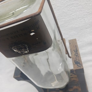 May include: A clear glass bottle with a brown label and a metal frame, displayed on a wooden stand. The label has text that is partially visible. The stand is black with a distressed finish.