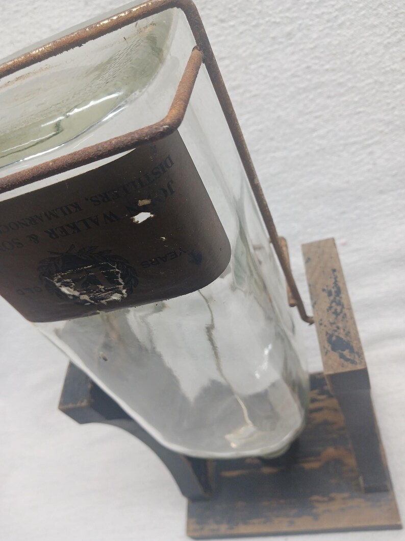 May include: A clear glass bottle with a brown label and a metal frame, displayed on a wooden stand. The label has text that is partially visible. The stand is black with a distressed finish.