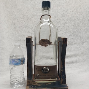 May include: A clear glass bottle with a black cap, held in a wooden stand. The stand is dark blue with a distressed finish and the words "Johnnie Walker" are visible. A plastic water bottle is next to the stand.