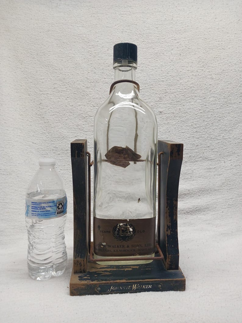 May include: A clear glass bottle with a black cap, held in a wooden stand. The stand is dark blue with a distressed finish and the words "Johnnie Walker" are visible. A plastic water bottle is next to the stand.