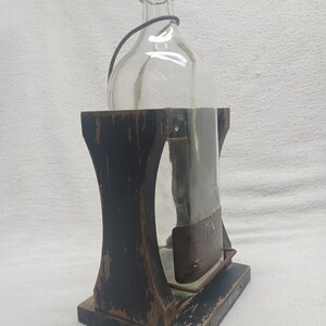 May include: A clear glass bottle with a black cap and a wire handle, held in a distressed black wooden stand. The stand has a rectangular base and a curved design. The bottle is partially encased in a metal sleeve.