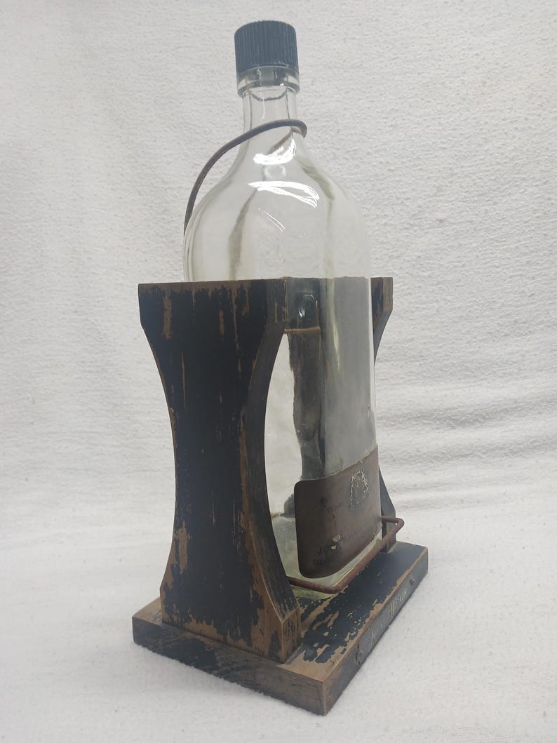 May include: A clear glass bottle with a black cap and a wire handle, held in a distressed black wooden stand. The stand has a rectangular base and a curved design. The bottle is partially encased in a metal sleeve.