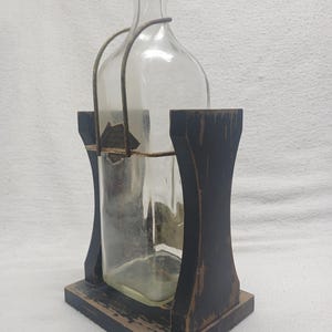 May include: A clear glass bottle with a black cap and a metal handle, held within a distressed black wooden holder. The holder has a rectangular base and arched sides, creating a rustic aesthetic.