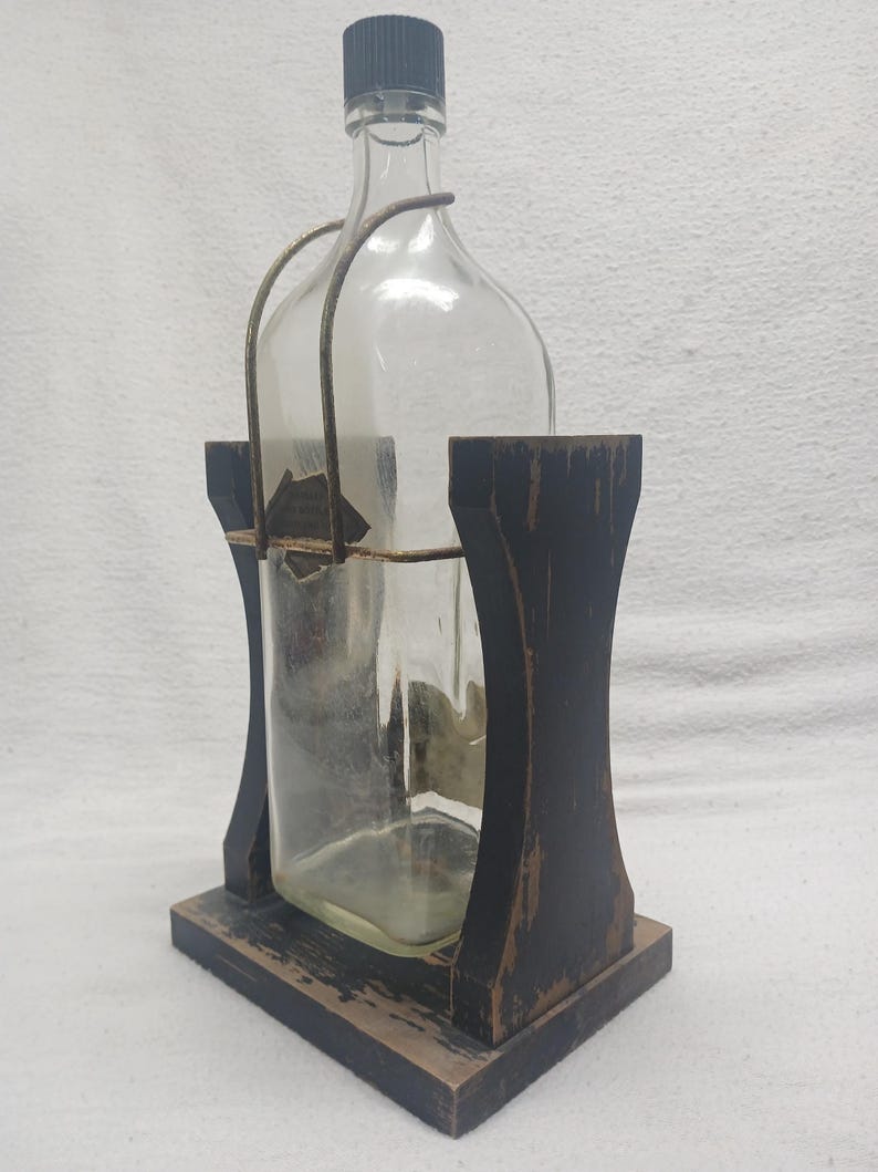 May include: A clear glass bottle with a black cap and a metal handle, held within a distressed black wooden holder. The holder has a rectangular base and arched sides, creating a rustic aesthetic.