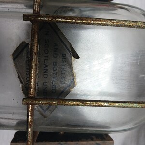 May include: A clear glass bottle with a label that reads "DISTILLED AND BOTTLED IN SCOTLAND UNDER SUPERVISION". The bottle is encased in a rusty metal cage.