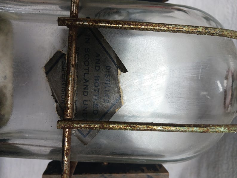 May include: A clear glass bottle with a label that reads "DISTILLED AND BOTTLED IN SCOTLAND UNDER SUPERVISION". The bottle is encased in a rusty metal cage.