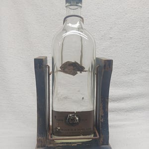 May include: A clear glass bottle with a black cap, held in a wooden stand. The bottle has a brown label with text. The stand is dark blue with a metal frame. The base of the stand reads "Johnnie Walker".