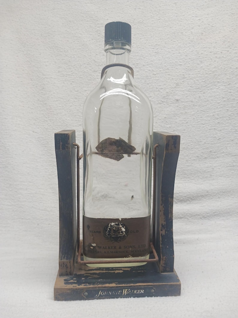 May include: A clear glass bottle with a black cap, held in a wooden stand. The bottle has a brown label with text. The stand is dark blue with a metal frame. The base of the stand reads "Johnnie Walker".