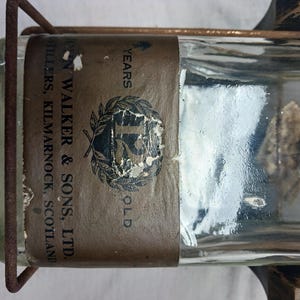 May include: A clear glass bottle with a brown label that reads "John Walker & Sons, Ltd. Distillers, Kilmarnock, Scotland" and "15 Years Old." The bottle is held in a metal cage.