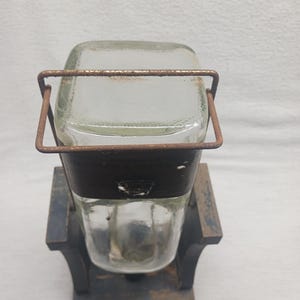 May include: A vintage glass jar with a rectangular top and a rounded bottom, held in place by a rusty metal frame. The jar sits on a dark wooden base. The design suggests a rustic, antique aesthetic.